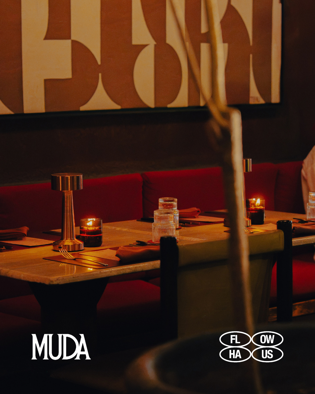 Candlelit tables and red banquettes inside MUDA, with the MUDA and FLOW HAUS PROJECT logos